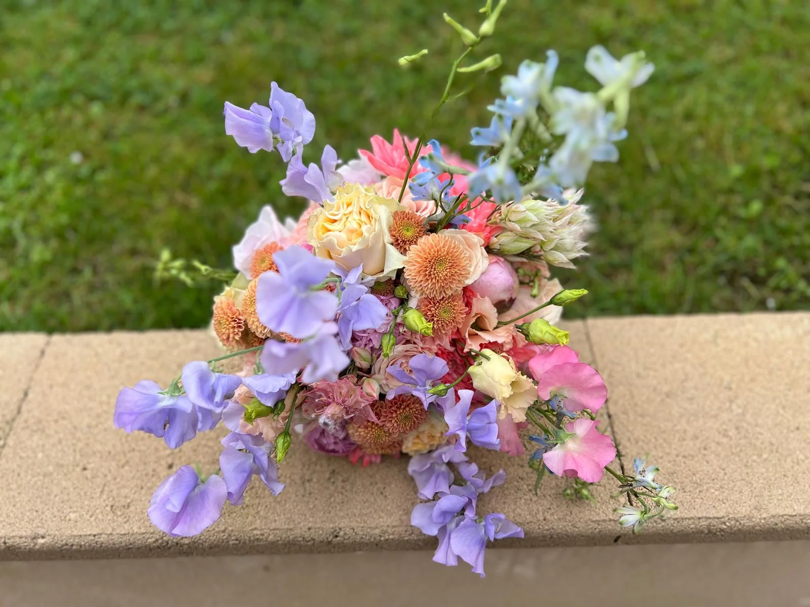 Bouquet de mariée pastel aux pois de senteur mauves, roses pêche et dahlias corail, posé sur un muret – Armony's Déco