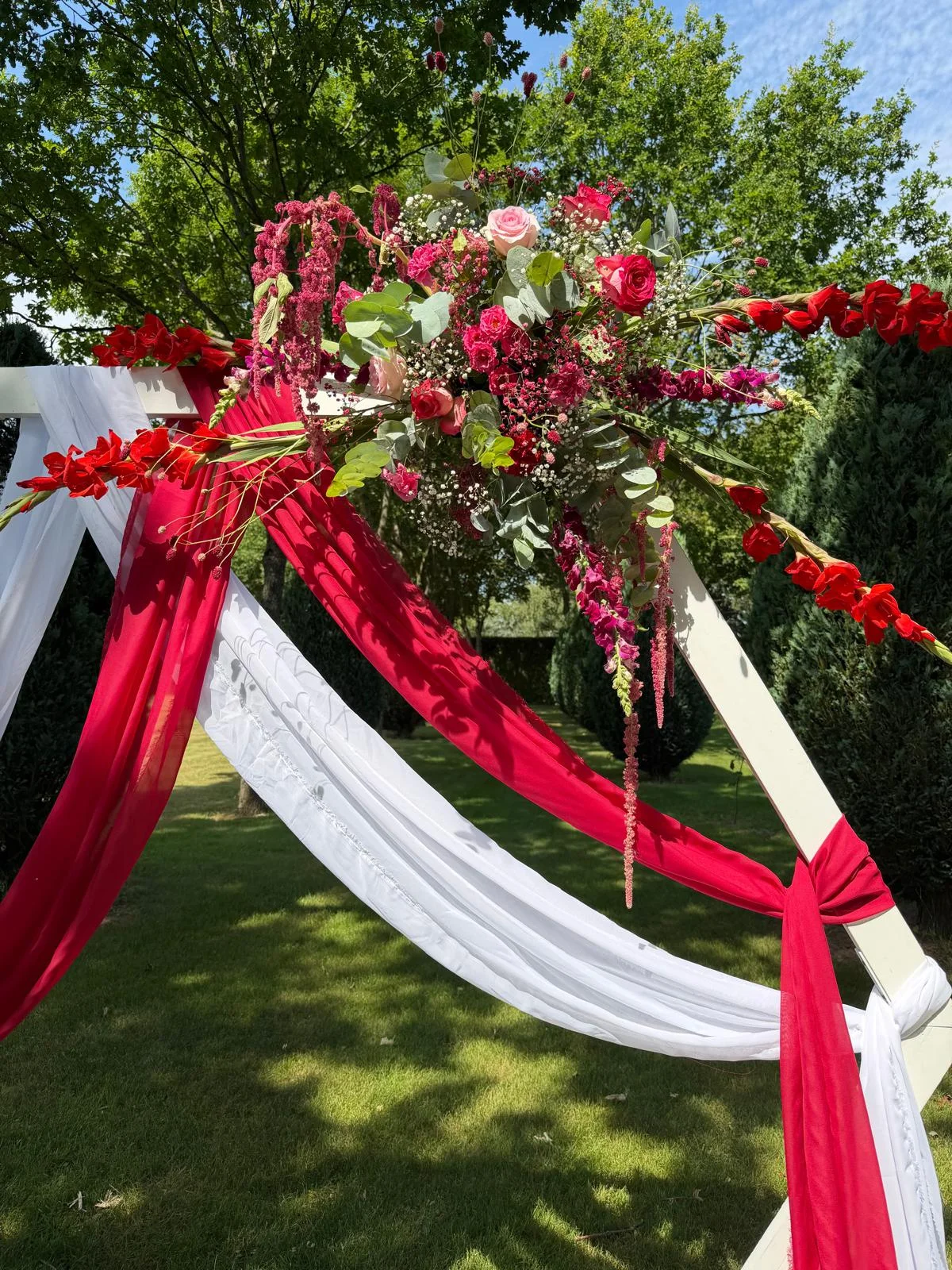 Arche aux voilages rouge et blanc fleurie de roses rouges, dans un jardin – Armony's Déco