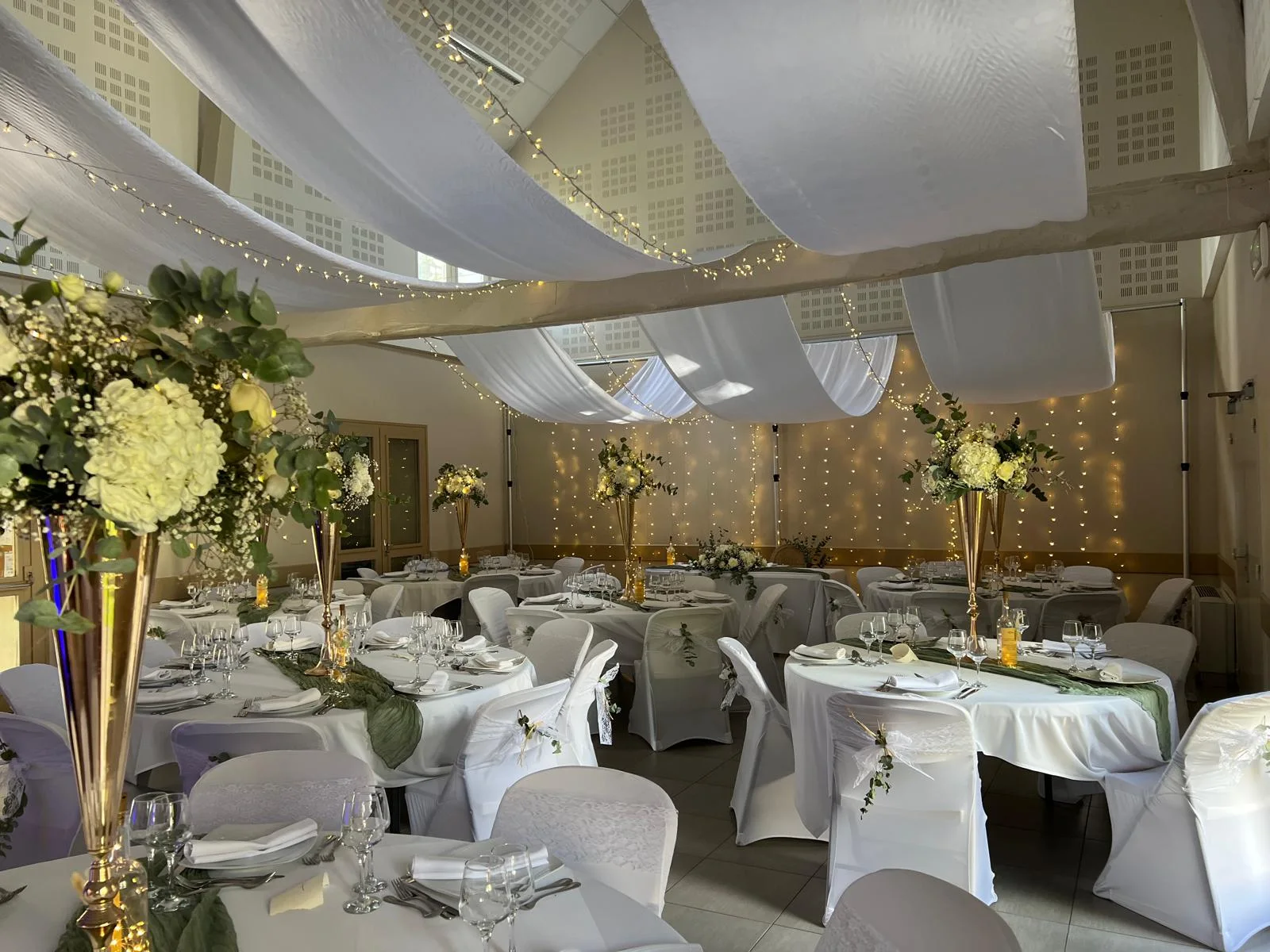 Salle de réception aux voilages blancs et guirlandes lumineuses, vases flûtes dorés fleuris – Armony's Déco