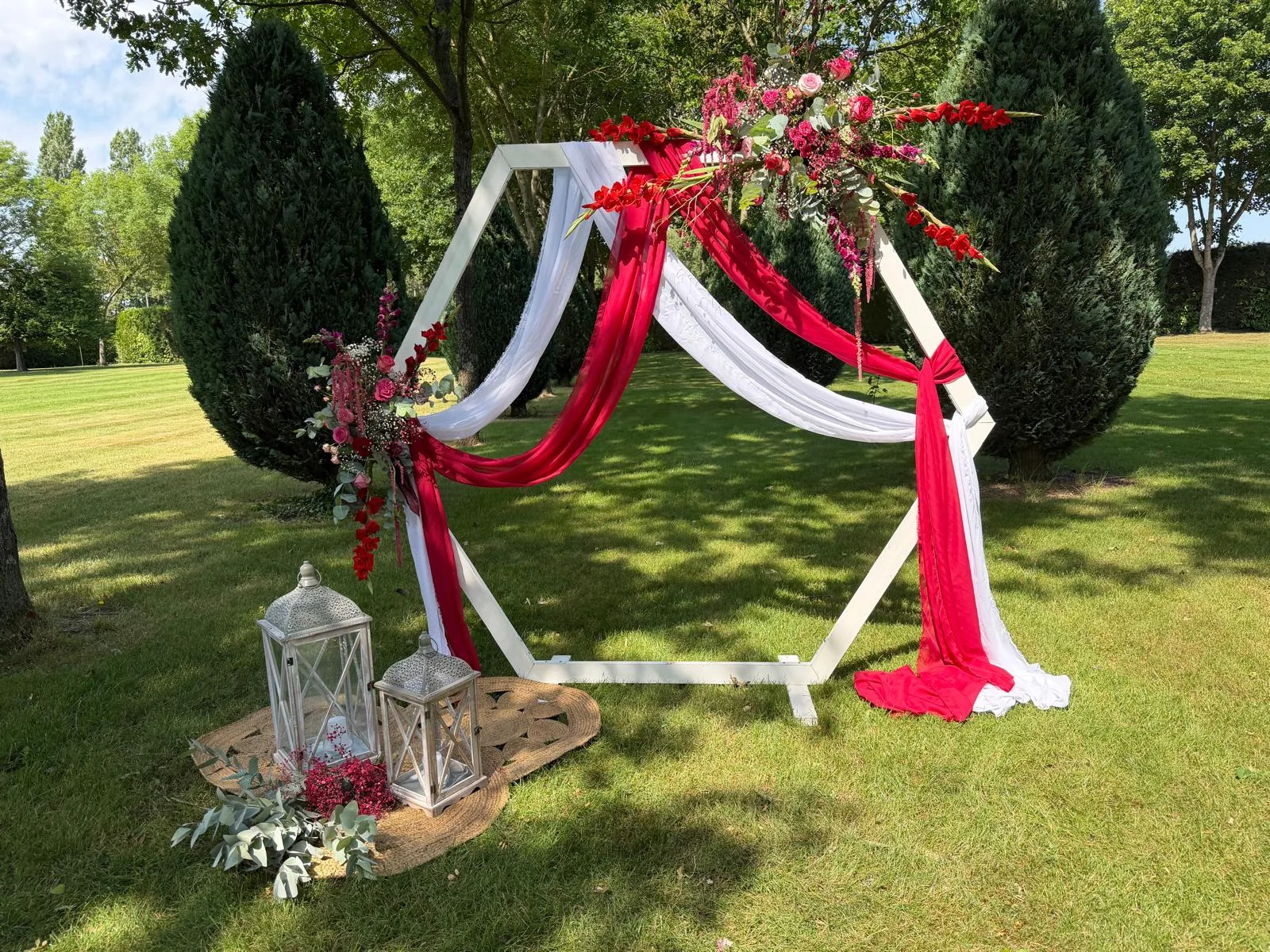 Arche hexagonale aux voilages blanc et rouge et lanternes, dans un parc – Armony's Déco