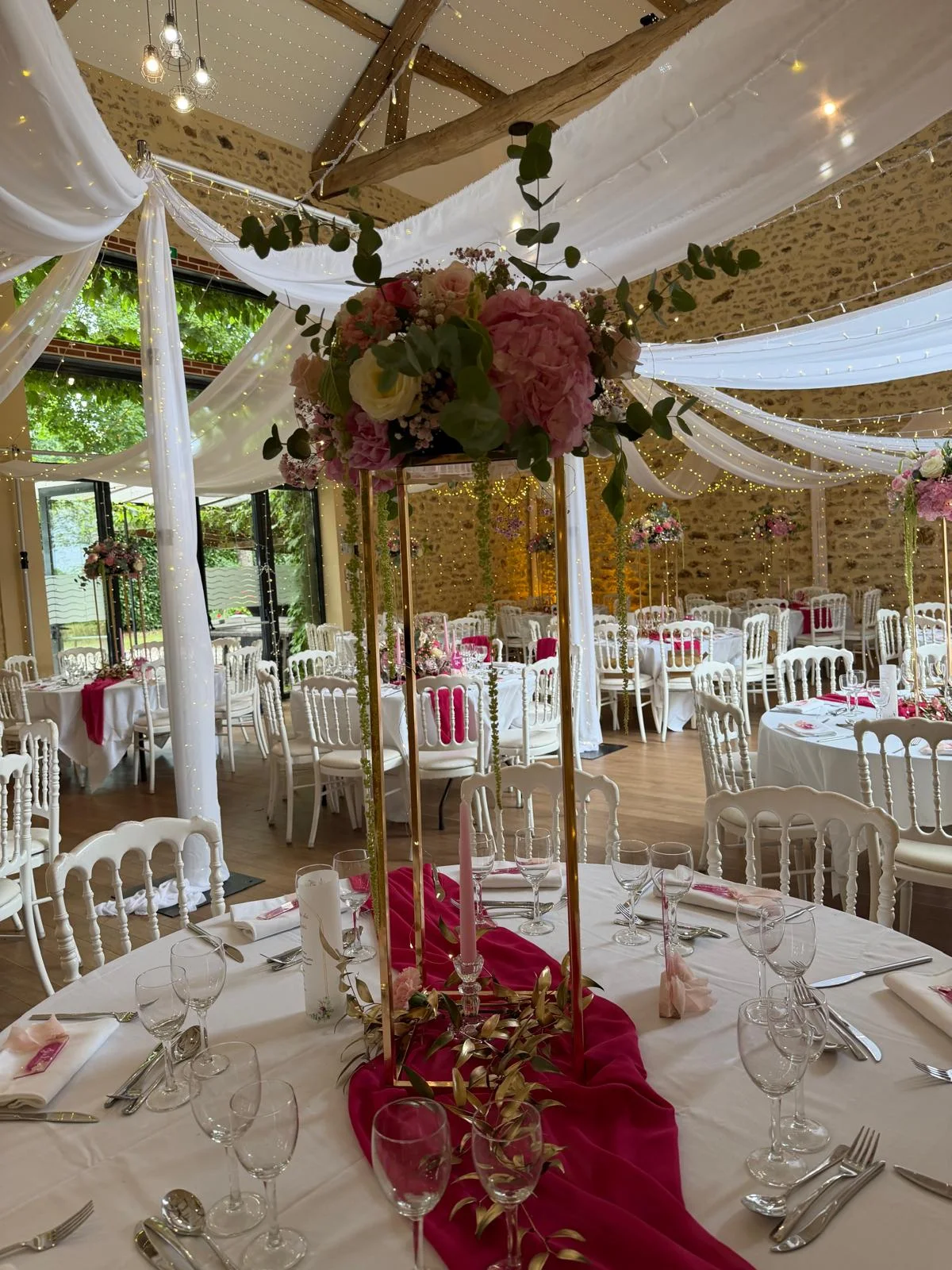 Centre de table haut sur pied doré, fleurs roses et bordeaux, voilages blancs – Armony's Déco